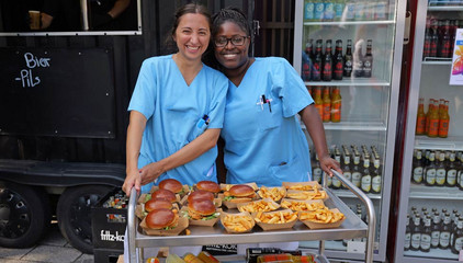 Zwei Mitarbeiterinnen in blauer Funktionskleidung stehen hinter einem Servierwagen mit frischen Burgern und Pommes und blicken fröhlich in die Kamera beim Albertinen-Sommerfest für die Mitarbeitenden aus den unterschiedlichen Einrichtungen der Immanuel Albertinen Diakonie am Standort Schnelsen auf dem Platz vor der Kirche am Albertinen Krankenhaus. Zwei Mitarbeiterinnen in blauer Funktionskleidung stehen hinter einem Servierwagen mit frischen Burgern und Pommes und blicken fröhlich in die Kamera beim Albertinen-Sommerfest für die Mitarbeitenden aus den unterschiedlichen Einrichtungen der Immanuel Albertinen Diakonie am Standort Schnelsen auf dem Platz vor der Kirche am Albertinen Krankenhaus.